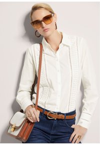 Witte knoop-overhemd met intricaten kantdetails, gecombineerd met blauwe jeans en een bruine riem. Accessoires omvatten een bruine tas en oversized zonnebril.