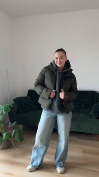 Oļivzaļš pūkains jaka ar kapuci, valkāta pāri pelēkai sporta džemperim. Brīvi pieguļoši gaiši zili džinsi un bēšas sporta kurpes. Telpās, ar zaļu dīvānu fonā.