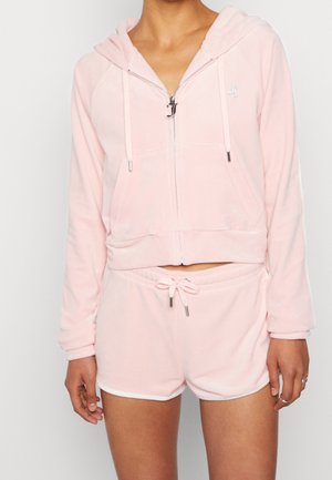 Person wearing light pink zip-up hoodie with front pockets and matching drawstring shorts, standing against a plain white background.