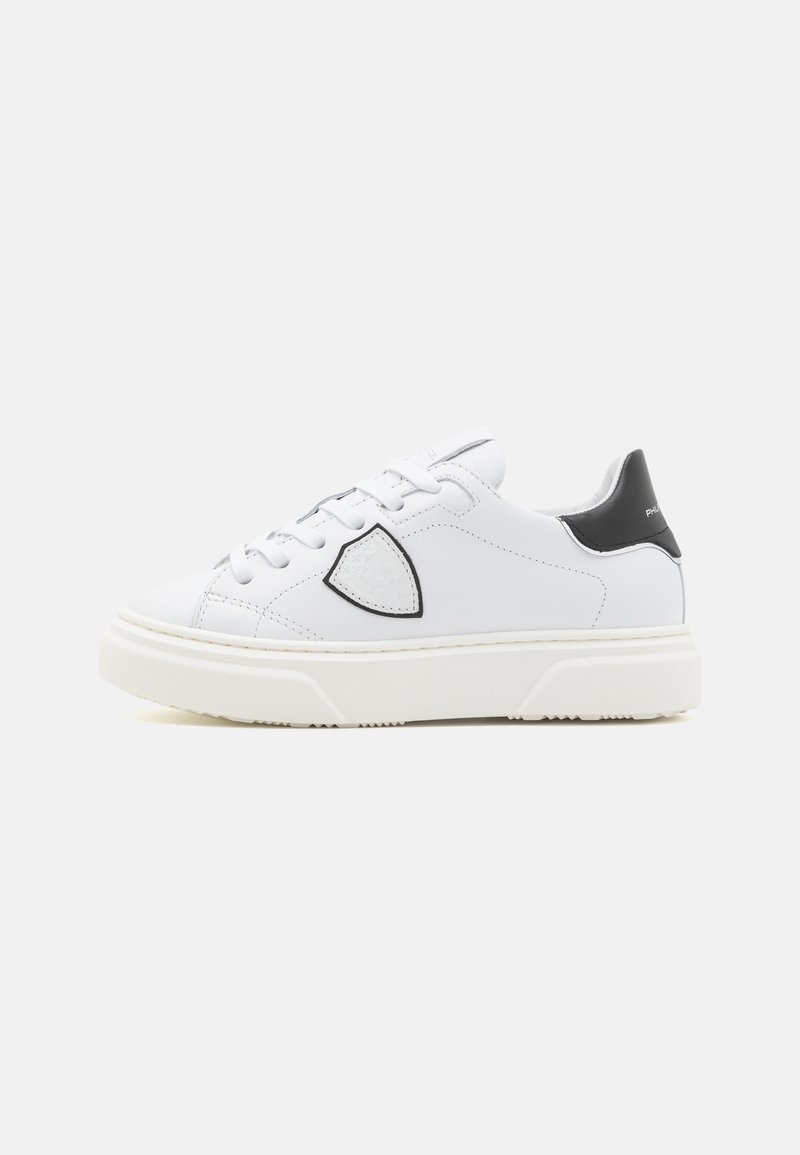 Sneakers en cuir blanc avec une semelle en caoutchouc texturée blanche, des accents de talon noirs et un contour décoratif noir autour du logo sur le côté.