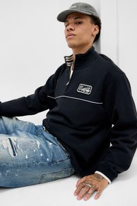 Ung mand med mønstret kasket, sort zip-up sweatshirt og slidte blå jeans, der sidder op ad en hvid væg, og viser håndtattooer og ringe.