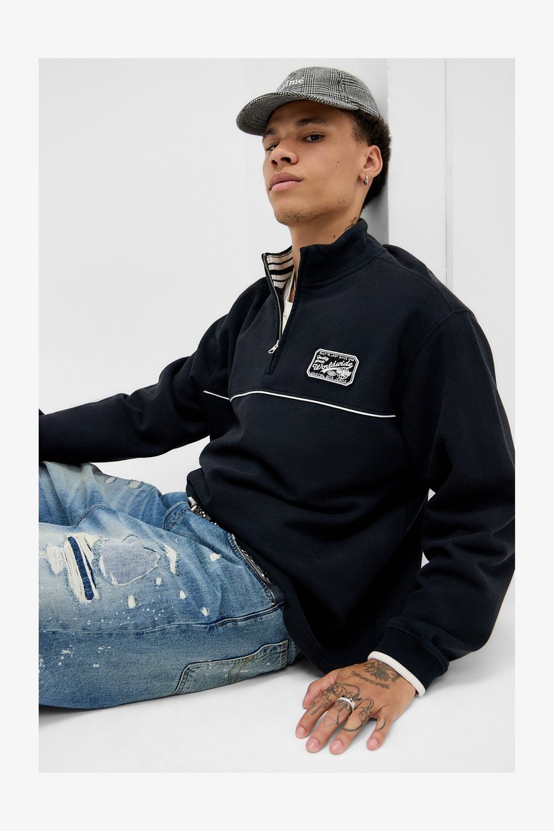 Ung mand med mønstret kasket, sort zip-up sweatshirt og slidte blå jeans, der sidder op ad en hvid væg, og viser håndtattooer og ringe.
