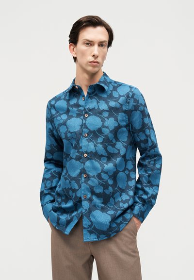 Νεαρός άνδρας με σκούρα μαλλιά φοράει μπλε πουκάμισο με λουλουδάτο μοτίβο και μπεζ παντελόνι, στέκεται με τα χέρια στις τσέπες μπροστά σε απλό φόντο.