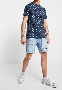 Camisa de manga corta azul marino con patrón blanco, combinada con pantalones cortos de mezclilla azul claro con desgaste y zapatillas altas blancas.
