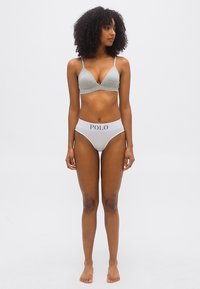 Γυναίκα όρθια ξυπόλυτη μπροστά σε λευκό φόντο, φοράει γκρι τρίγωνο σουτιέν και λευκά εσώρουχα με την επιγραφή "POLO" στη μέση.