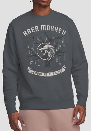 Man wearing dark gray sweatshirt with "Kaer Morhen School of the Wolf" text and a wolf head and tree graphic on the chest.