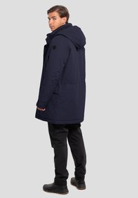 Parka imbottita blu navy con cappuccio, dotata di cordino in vita, tasche laterali e dettagli neri. Abbinata a pantaloni neri e stivali.