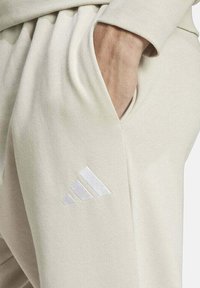 Pantaloni della tuta beige con una tasca laterale e logo Adidas bianco con tre strisce diagonali. Tessuto morbido e texture lavorata.