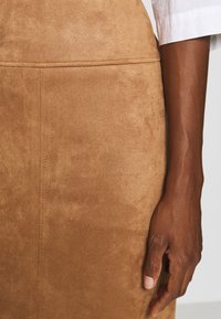 Jupe en suède marron avec une texture lisse, présentant un design simple et des coutures soignées. Une main est posée sur la surface de la jupe.