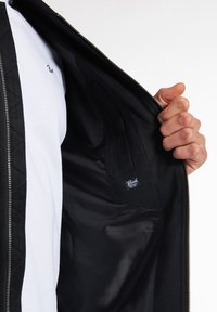 Jachetă neagră cu textură matlasată, închidere cu fermoar și un interior negru asemănător satinului. Tricou alb vizibil dedesubt cu un mic detaliu logo.