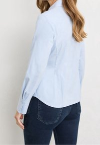 Light blue fitted shirt with long sleeves, featuring buttoned cuffs and a smooth texture. Paired with dark blue jeans, shown from the back.