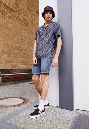 Chemise à manches courtes noir et grise à motif damier, short en denim, chaussures noires à enfiler avec des détails de flammes, et chaussettes blanches mi-mollet.