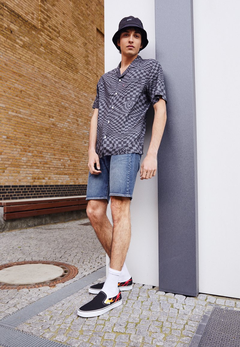 Camisa de cuadros en blanco y negro de manga corta, pantalones cortos de mezclilla, zapatos negro sin cordones con detalles de llamas y calcetines blancos.