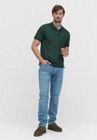 Grön polo med klassisk krage, med tre knappar. Bärs tillsammans med ljusblå jeans och bruna skor. Enfärgad bakgrund.