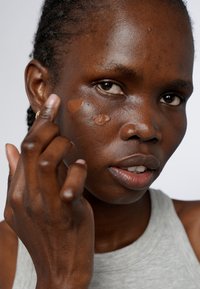 Le teint applique un produit fluide sur la joue, le produit étant visible sous forme de gouttes lisses et marron clair contre la peau. Vue en gros plan.