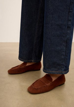Mocassins marron en daim sans lacets portés avec un jean large bleu foncé sur un sol clair.