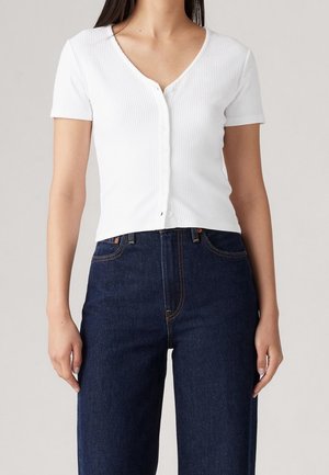 Mujer con una camiseta blanca de manga corta con botones y textura acanalada, y vaqueros azul oscuro de tiro alto, sobre un fondo liso.