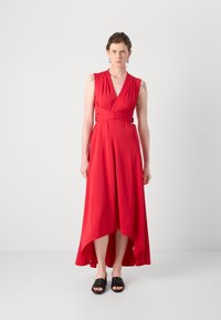 Robe sans manches rouge avec un décolleté en V profond, taille cintrée avec des accents métalliques et un ourlet asymétrique. Portée avec des chaussures à glissière noires.