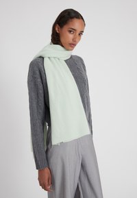 Sciarpa verde menta chiaro avvolta attorno al collo, realizzata in un tessuto morbido, abbinata a un maglione grigio testurizzato e pantaloni grigio chiaro.