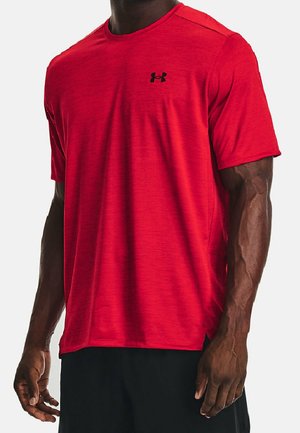 Mand iført en rød kortærmet atletisk skjorte med et lille sort Under Armour-logo på brystet og sorte shorts.