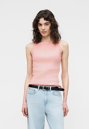 Kvinde med krøllet mørkt hår iført ærmeløs lyserød top, lyseblå jeans og sort bælte, stående mod en ensfarvet lys baggrund.