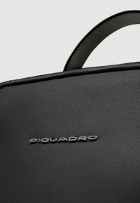 Zwarte leren tas met een gladde textuur, ritssluiting en een opvallend Piquadro-logo in metalen letters op het oppervlak.