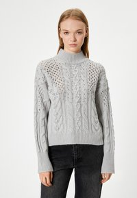 Pull en tricot gris clair avec un col haut, des motifs en câble élaborés, et une coupe décontractée avec des poignets et un ourlet côtelés.