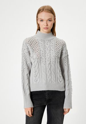 Pull en tricot gris clair avec un col haut, des motifs en câble élaborés, et une coupe décontractée avec des poignets et un ourlet côtelés.