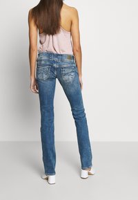 Jeans en denim bleu skinny avec poches arrière ornées de surpiqûres décoratives. Portés avec un haut sans manches rose clair et des chaussures à talons blanches.