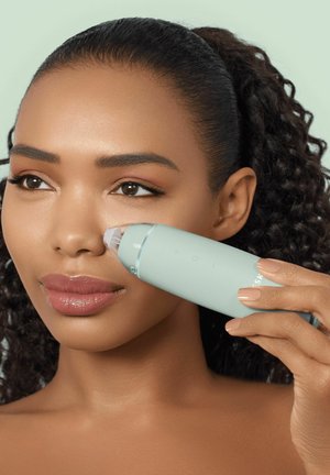 Woman using a handheld facial pore vacuum on her right cheek, with curly hair pulled back and natural makeup.