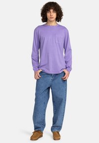 Langärmliges lavendelfarbenes Shirt mit einer Brusttasche, kombiniert mit locker sitzenden blauen Jeans und braunen Wildlederschuhen. Lockige Haare umrahmen das Gesicht.