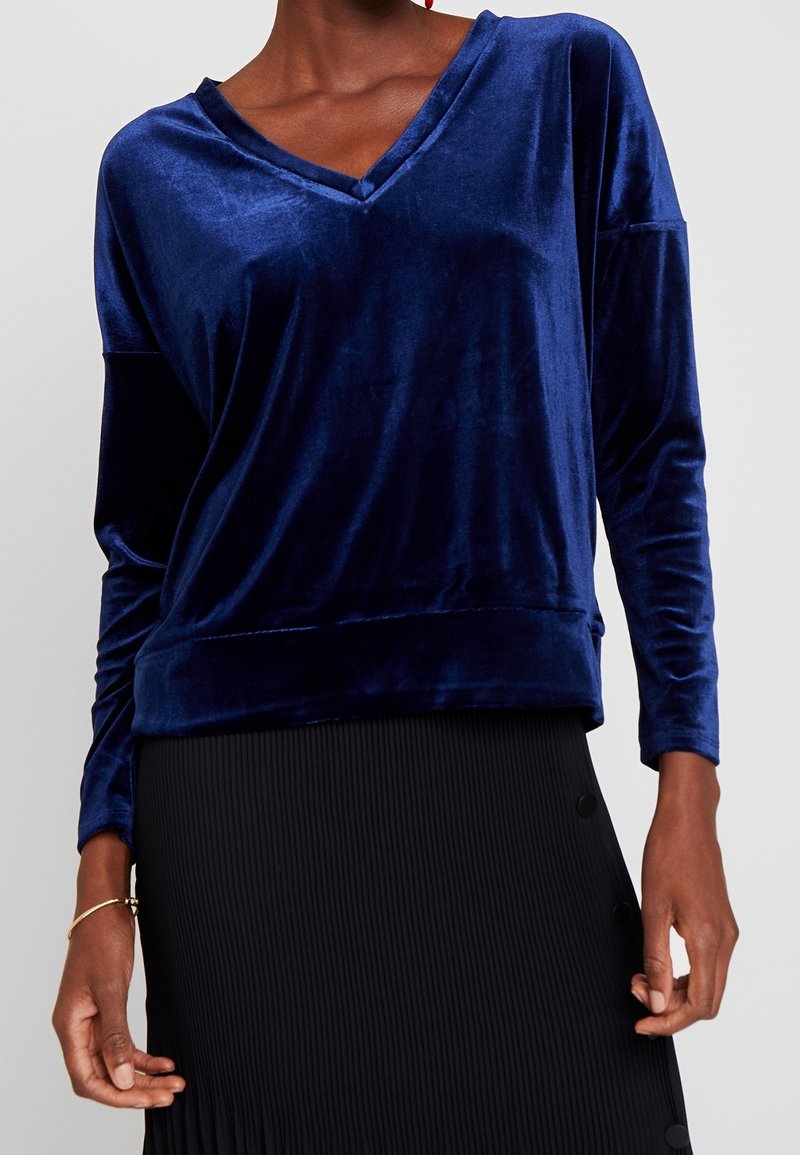 Top a maniche lunghe in velluto blu navy con scollo a V, caratterizzato da una vestibilità rilassata e una texture morbida, abbinato a una gonna nera plissettata.