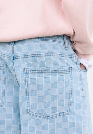 Pantaloni scurți din denim albastru deschis cu carouri, purtați cu un pulover tricotat roz pal băgat în pantaloni, mâna parțial în buzunarul lateral.