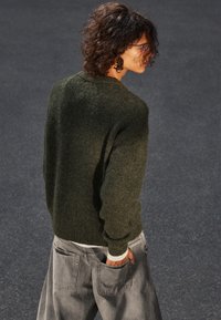 Dunkelgrüner Strickpullover in lockerer Passform, mit Raglanärmeln und gerippten Bündchen, kombiniert mit hellgrauen, lässigen Jeans mit großen Taschen.