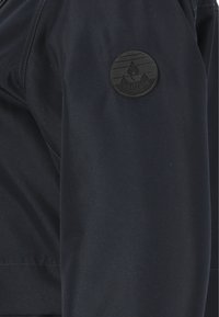 Mörkblå jacka med slät väv, en cirkulär svart patch med bergdesign och texten "Whistler Danmark" på vänster ärm.