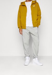 Sudadera amarilla con cremallera, camiseta blanca, pantalones de chándal grises con logo y zapatillas deportivas blancas. Tejido suave, ajuste casual y diseño con capucha.