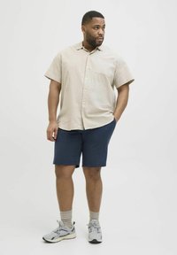 Beige Kurzarmhemd mit Knopfleiste und strukturiertem Muster, kombiniert mit marineblauen Shorts. Graue Sportschuhe und beige Socken runden das Outfit ab.