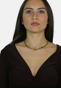 Złoty naszyjnik z okrągłym wisiorkiem, z przeplatanymi ogniwami. Nosi się go z ciemnym, głębokim dekoltem w kształcie V. Gładka, polerowana faktura.