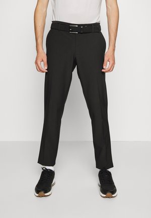 Homme portant un pantalon de costume noir avec une ceinture tissée noire, des chaussettes blanches et des baskets noires, debout devant un fond uni.