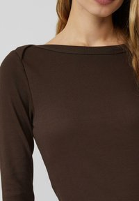Brązowa prążkowana bluzka z długim rękawem, z szerokim dekoltem i gładką fakturą. Zbliżenie na detal tkaniny i szew na ramieniu.
