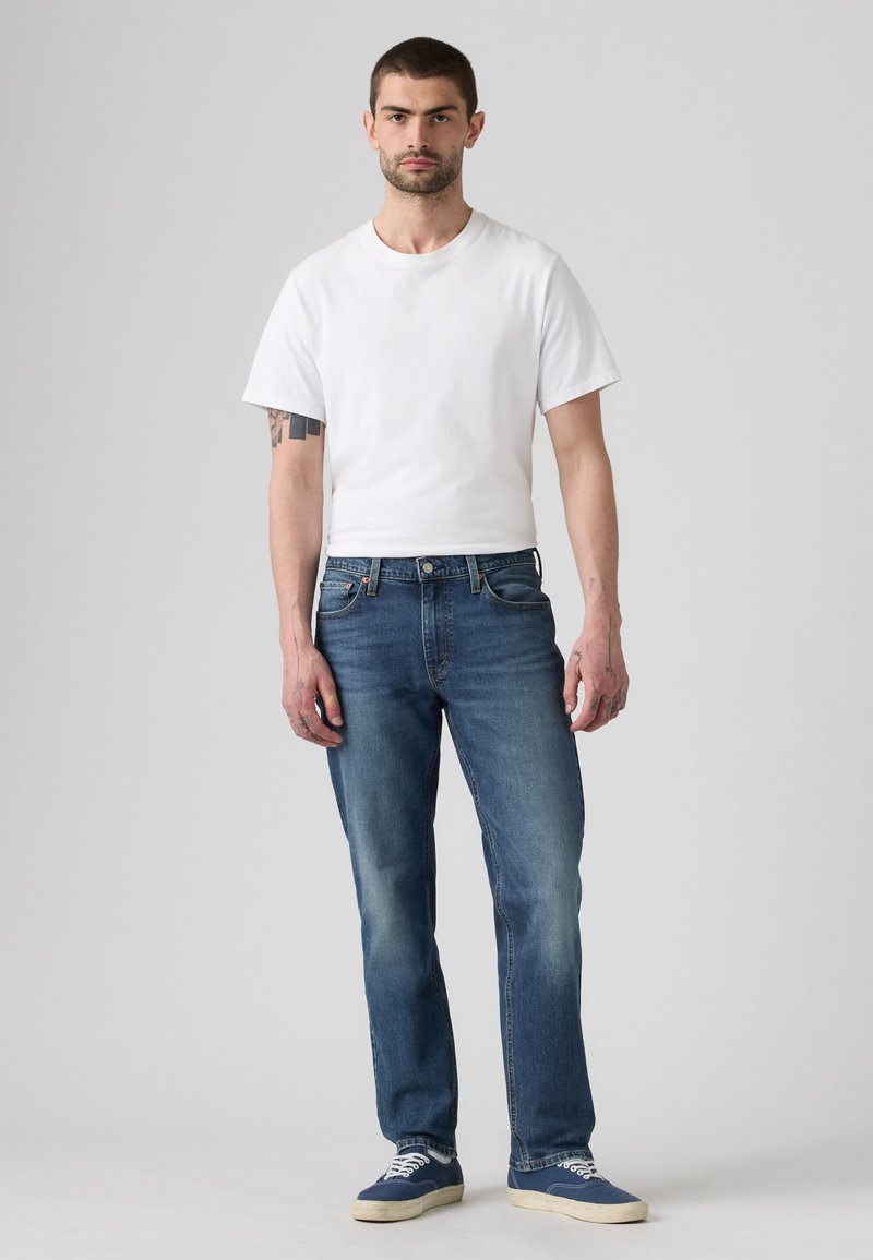 Man standing wearing a plain white short-sleeve t-shirt, blue jeans, and blue sneakers with white soles on a gray background.