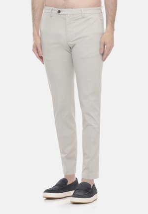 Pantaloni beige slim fit con passanti per cintura, chiusura con bottone e zip indossati da una persona abbinati a scarpe slip-on nere con suole bianche.