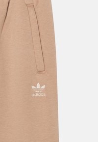 Béžové tepláky vyrobené z měkké tkaniny, s bočními kapsami a bílým vyšitým logem Adidas na levé noze.