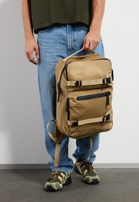 Rucsac din nailon bej cu compartimente cu fermoar, accente hardware negre și bretele ajustabile, ținut de o persoană îmbrăcată lejer.
