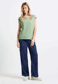 Zielona bawełniana koszulka z krótkim rękawem i okrągłym dekoltem, w połączeniu z luźnymi ciemnoniebieskimi dżinsami i beżowymi trampkami.