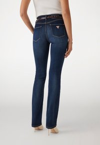 Dark blue, flared jeans with a mid-rise waist, featuring a stitched design on back pockets and a patterned belt with a pink logo.