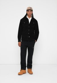 Veste en velours côtelé noir avec deux poches poitrine, fermeture à boutons, portée sur une chemise blanche. Un pantalon cargo noir et des bottes de travail beiges complètent la tenue.