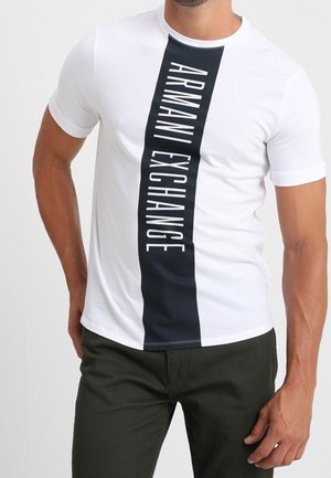 Mężczyzna w białej koszulce z pionowym czarnym paskiem i nadrukiem "ARMANI EXCHANGE" białymi literami na środku.