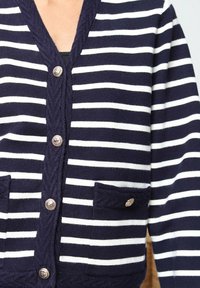 Cardigan rayé bleu marine et blanc avec un col en V, tricot texturé, deux poches avant et des boutons métalliques pour la fermeture.