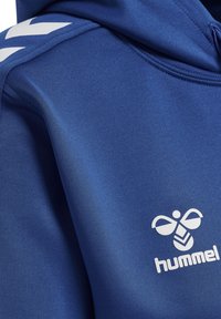Blå sports hættetrøje med hvidt hummel-logo og hvidt chevron-stribe på skuldersømmen.
